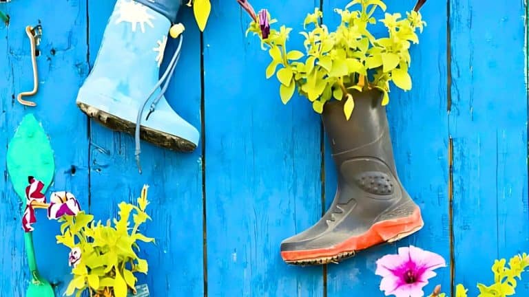 Two rain boots repurposed as planters with green plants, mounted on a blue wooden fence, accompanied by scattered pink and purple flowers.