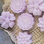 Six lavender-colored flower-shaped soap bars, inspired by whipped sugar scrub soap recipes, are arranged on a crocheted doily beside a metal surface. A small "Off Grid Life" logo appears in the lower right corner.