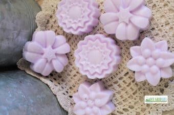 Six lavender-colored flower-shaped soap bars, inspired by whipped sugar scrub soap recipes, are arranged on a crocheted doily beside a metal surface. A small "Off Grid Life" logo appears in the lower right corner.