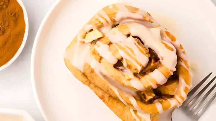 A cinnamon roll with icing on a white plate, partially eaten, next to a fork.