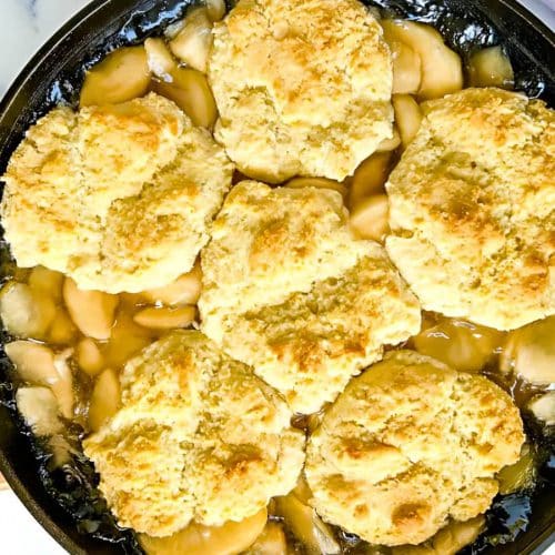 A skillet of apple cobbler with six biscuit toppings on a marble surface.
