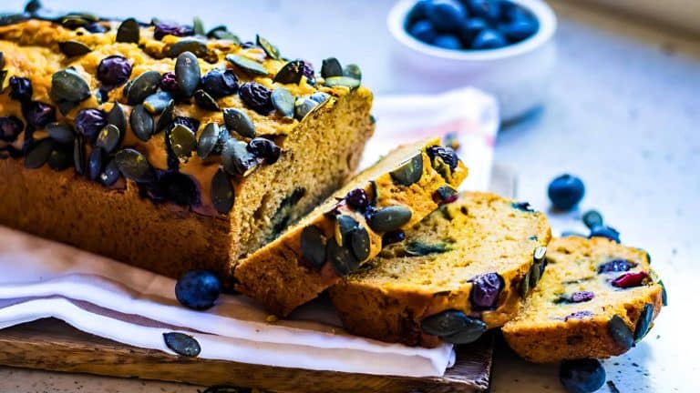 A sliced loaf of pumpkin bread topped with seeds and blueberries on a wooden board. A small bowl of blueberries is in the background.