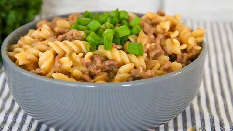 A bowl of fusilli pasta with ground beef, topped with chopped green onions, sits on a striped cloth.