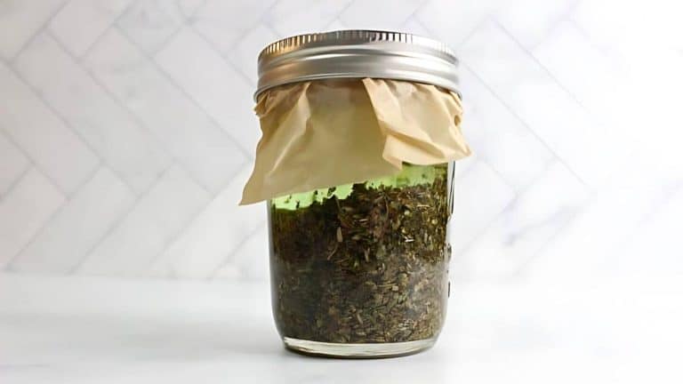 A jar filled with dried herbs, covered with paper and a metal ring lid, placed against a white tiled background.