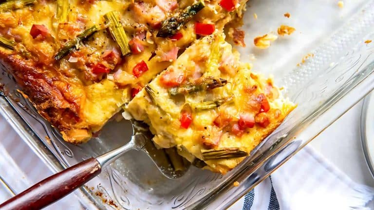 A slice of vegetable casserole with visible asparagus and red peppers is being served from a glass baking dish.