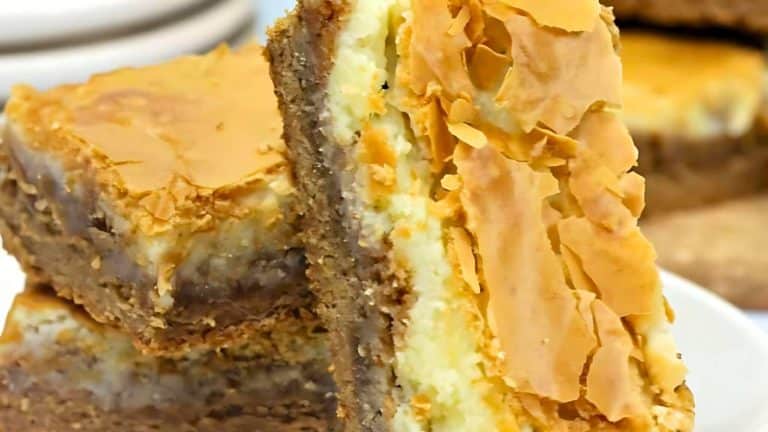 Close-up of layered dessert bars with a flaky top and creamy filling on a white plate.