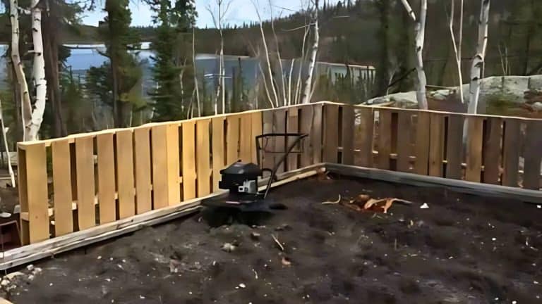 A garden tiller rests on dark soil in a wooden fenced area surrounded by trees, with a lake visible in the background.