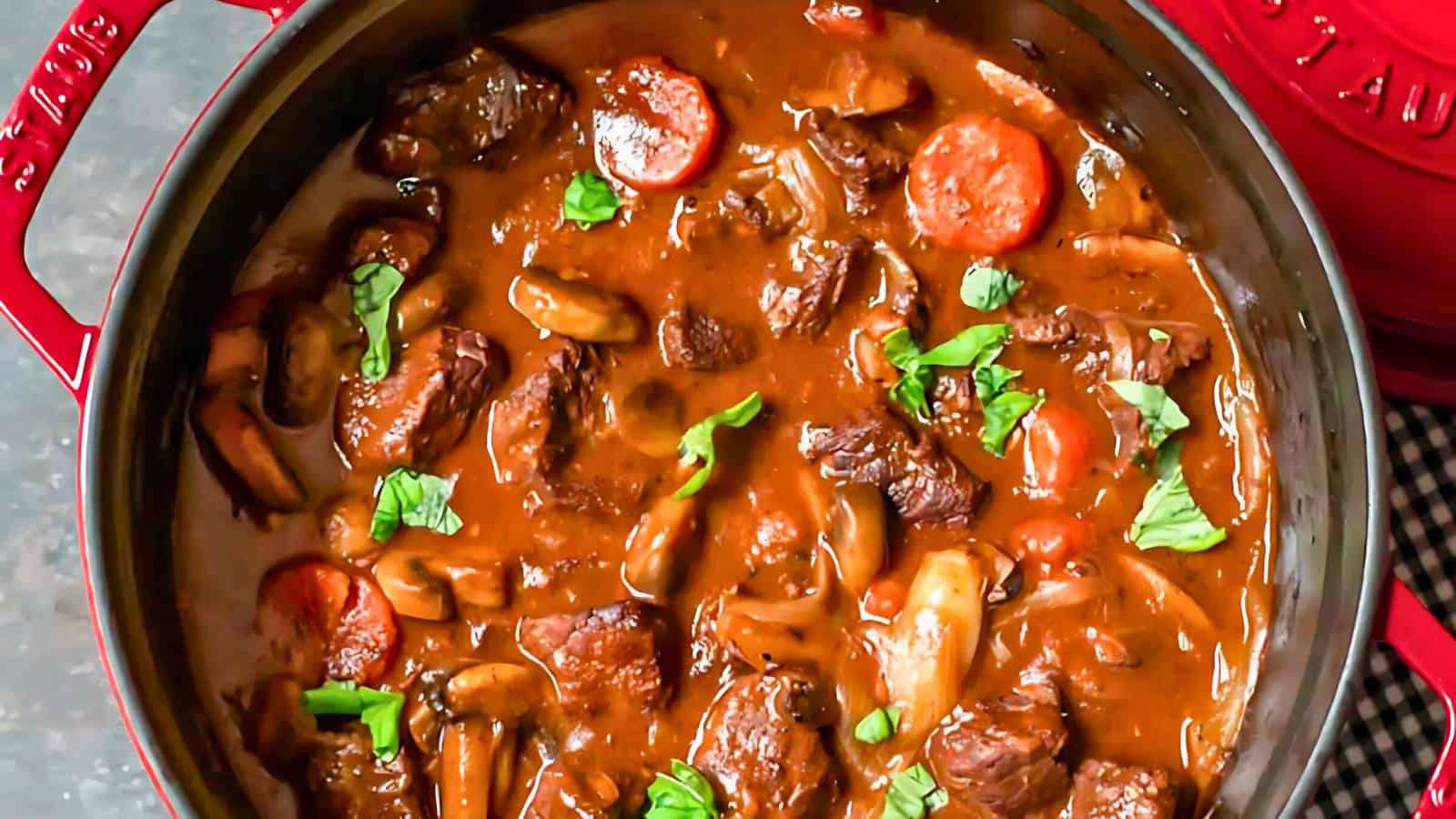 A pot of beef stew with carrots, mushrooms, and garnished with parsley in a red pot.