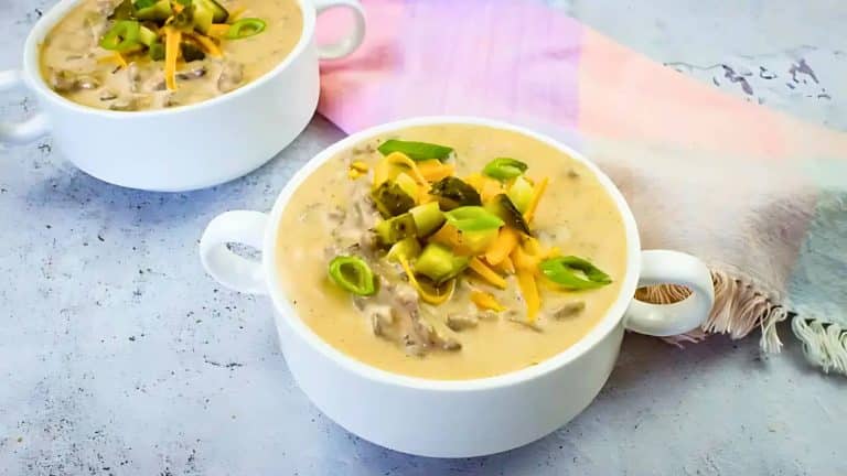 Two bowls of cheeseburger soup topped with chopped pickles, green onions, and shredded cheese. A colorful napkin is beside the bowls on a light-colored surface.