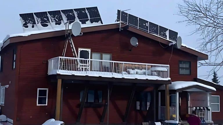 A snow-covered house with solar panels and satellite dishes on the roof, a ladder on the balcony, and snow on the ground and railing.