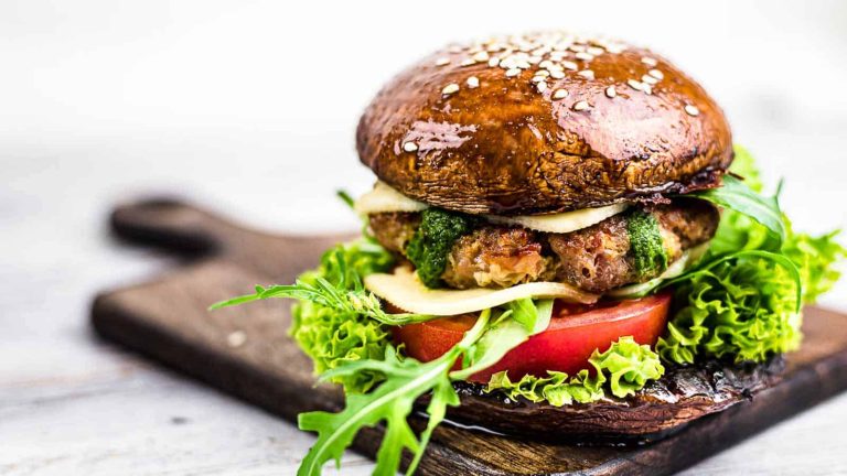 A gourmet burger made with a portobello mushroom bun, lettuce, tomato, cheese, a meat patty, and green sauce, served on a wooden board.