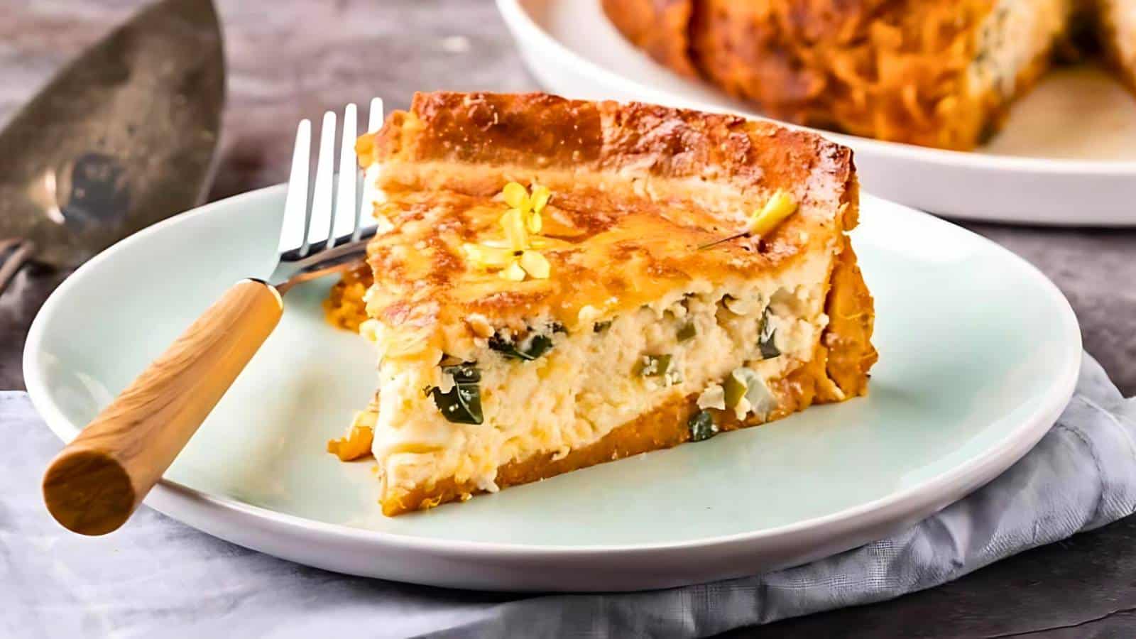 A slice of quiche is served on a white plate with a fork resting on the side. The quiche has a golden-brown crust and visible filling with cheese and herbs. A larger portion is in the background.