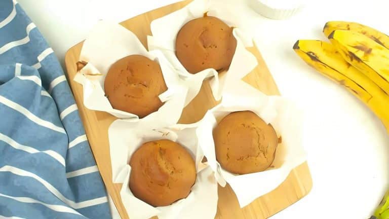 Four golden-brown muffins on parchment paper rest on a wooden board. A striped blue cloth is on the left, and two bananas are on the right.