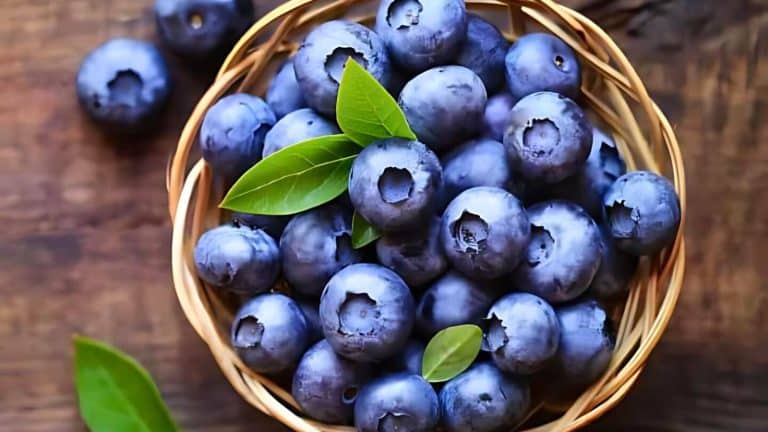 A woven basket filled with fresh blueberries and green leaves, placed on a wooden surface.