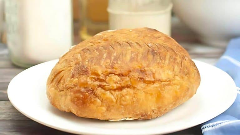 A round loaf of crusty bread sits on a white plate, with a blurred background showing kitchen items.