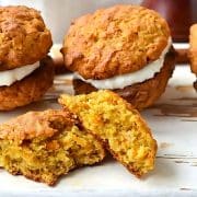 Four pumpkin cookies with cream filling are arranged on a wooden surface. One cookie is broken apart, revealing its texture. Anise stars are also visible.