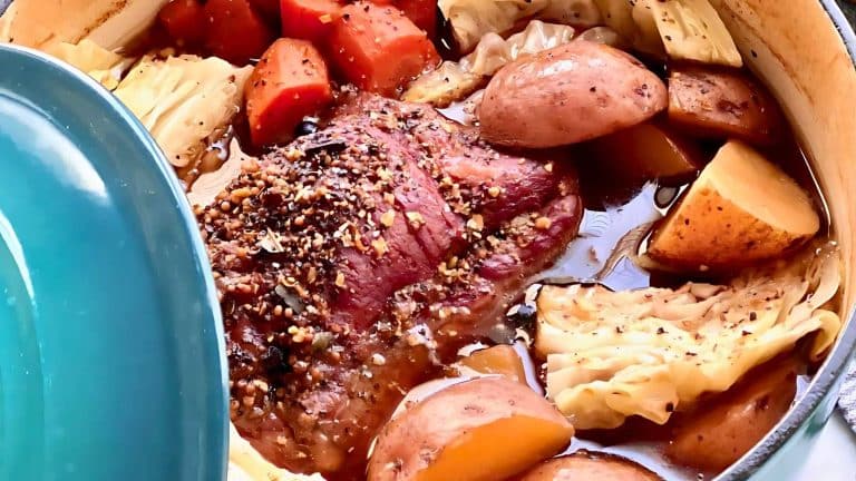 A close-up of a pot roast with seasoned beef, carrots, cabbage, and potatoes cooked in broth. A teal lid is partially open on the side of the pot.
