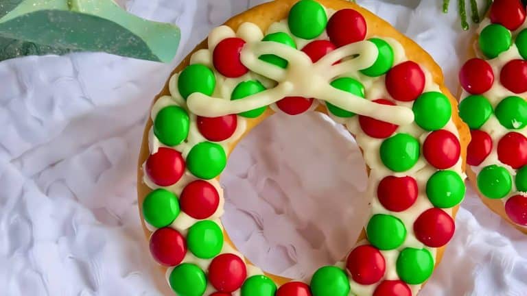 A donut decorated with red and green candy pieces and a white icing bow, resembling a festive wreath, placed on a white cloth.