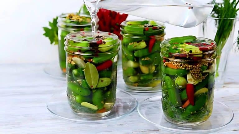 A person pours liquid into jars filled with pickling cucumbers, chili peppers, garlic, mustard seeds, and bay leaf on a white surface.