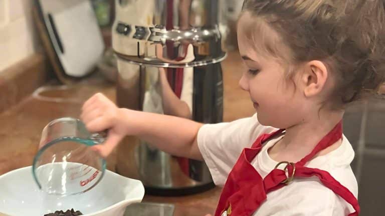 Child in a red apron pours ingredients from a measuring cup into a bowl on a kitchen counter.