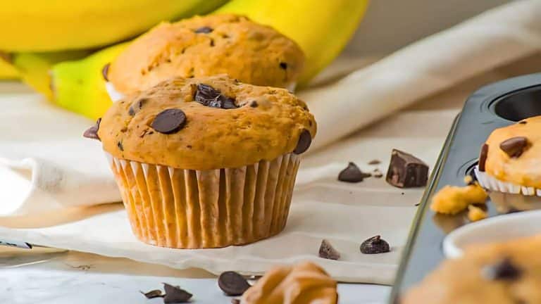 A chocolate chip muffin on a table with banana and chocolate pieces in the background, next to a muffin tin.