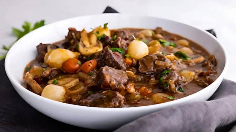 A white bowl filled with beef stew, containing chunks of beef, carrots, mushrooms, onions, and garnished with parsley, placed on a dark cloth.