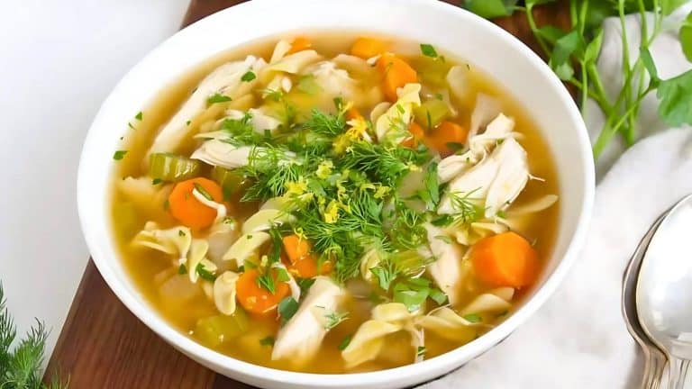 A bowl of chicken noodle soup with carrots, celery, and herbs, garnished with dill and lemon zest, placed on a wooden surface next to a spoon.