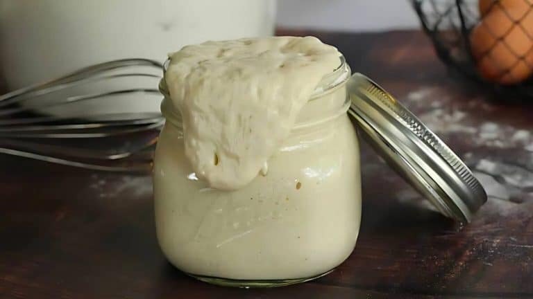 Glass jar filled with overflowing bread dough on a wooden surface, next to a metal whisk and a slightly opened lid.