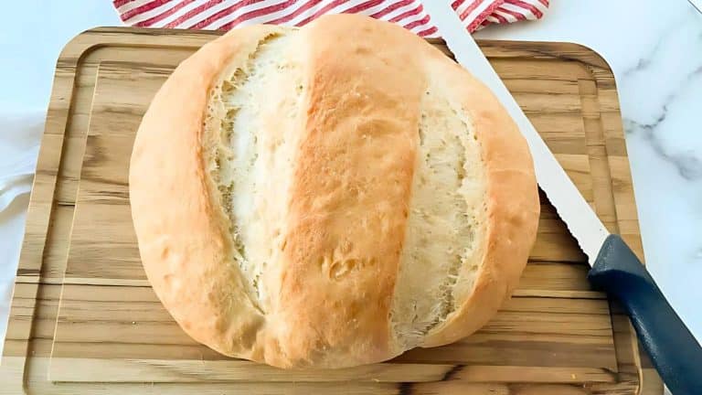 Round loaf of bread with two deep cuts, placed on a wooden cutting board beside a serrated knife.