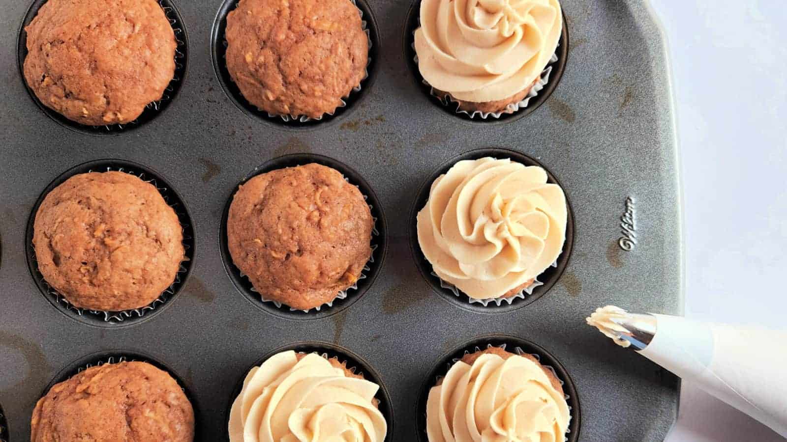 Cupcakes in a muffin tray, half plain and half with piped swirls of frosting. A piping bag is nearby.