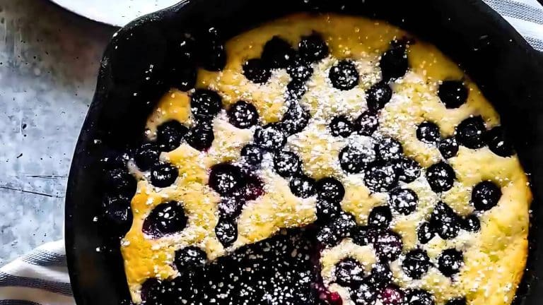 A blueberry clafoutis in a skillet with powdered sugar on top, a wedge missing.