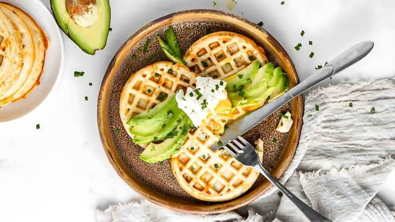 A plate with two waffles, sliced avocado, and a poached egg. A fork and knife are cutting into the egg. Avocado slices and chives are scattered around. A half avocado is visible in the background.