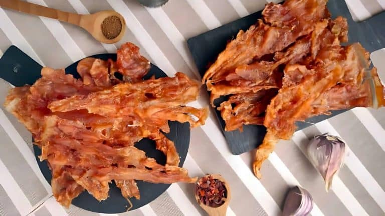 Two plates of crispy fried chicken skins on a striped tablecloth, with a wooden spoon of brown spice and cloves of garlic nearby.