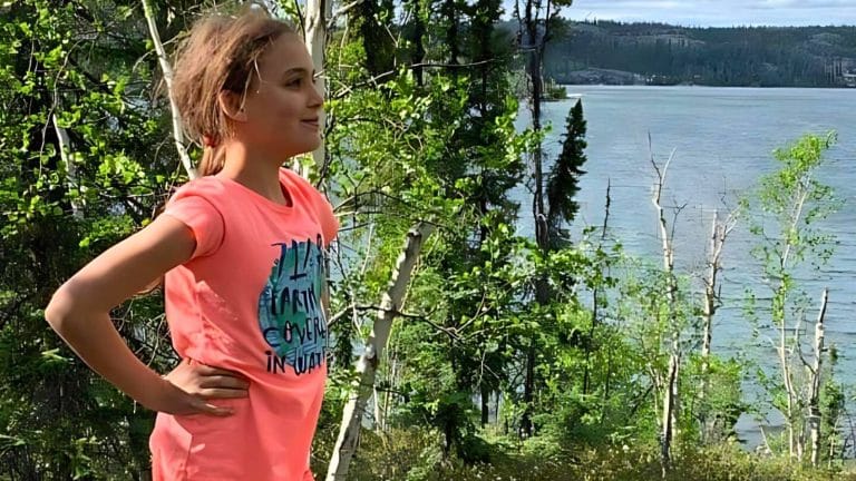 Person in a pink shirt stands with hands on hips, looking out at a scenic view of a forested area with a body of water and hills in the background.