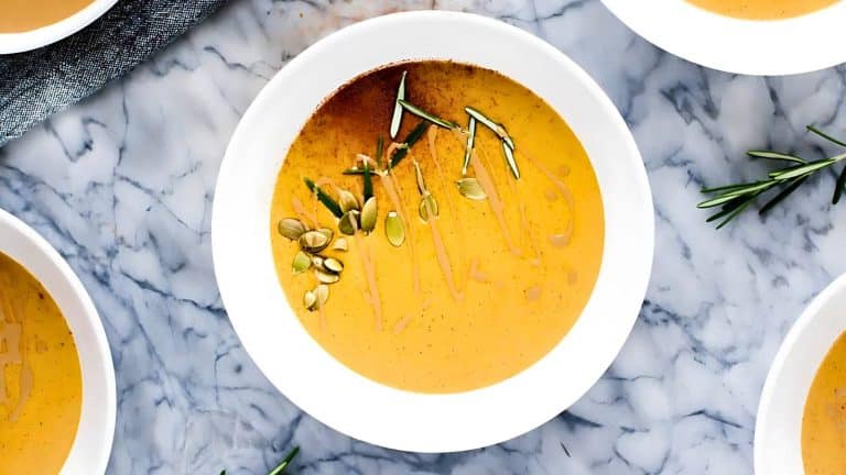 Bowl of soup garnished with pumpkin seeds and rosemary on a marble surface.