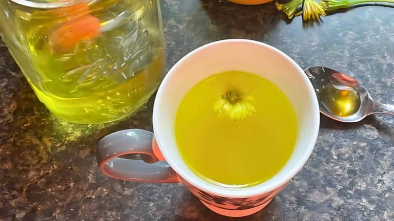 A white mug filled with yellow liquid and a flower on top, next to a spoon and a large jar of similar liquid on a dark surface.