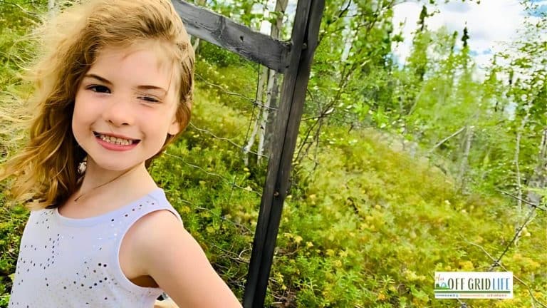 A young girl with light brown hair winking and smiling, standing outside near a wire fence, with green foliage in the background. A logo reading "OFF GRID LIFE" is in the bottom right corner.