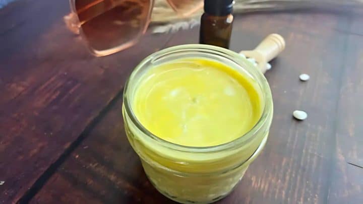 A glass jar filled with yellow cream on a wooden table, accompanied by a small bottle, sunglasses, and a honey dipper.