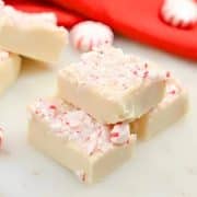 White chocolate peppermint fudge with crushed candy pieces on top, surrounded by whole peppermint candies and red fabric in the background.