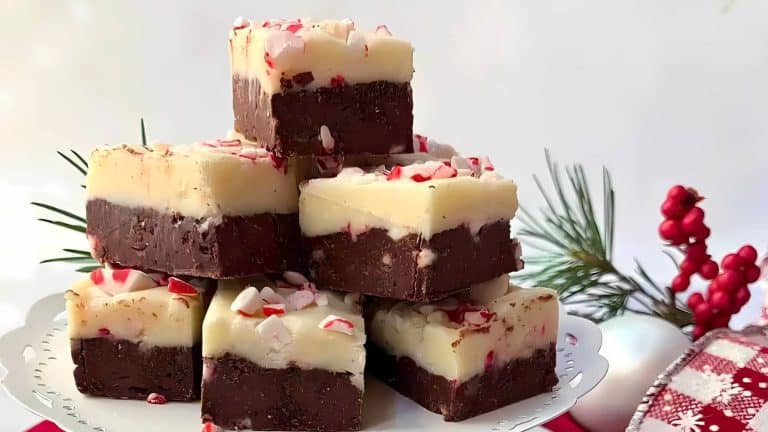 A stack of chocolate and white fudge squares topped with crushed peppermint, displayed on a white cake stand with festive greenery in the background.