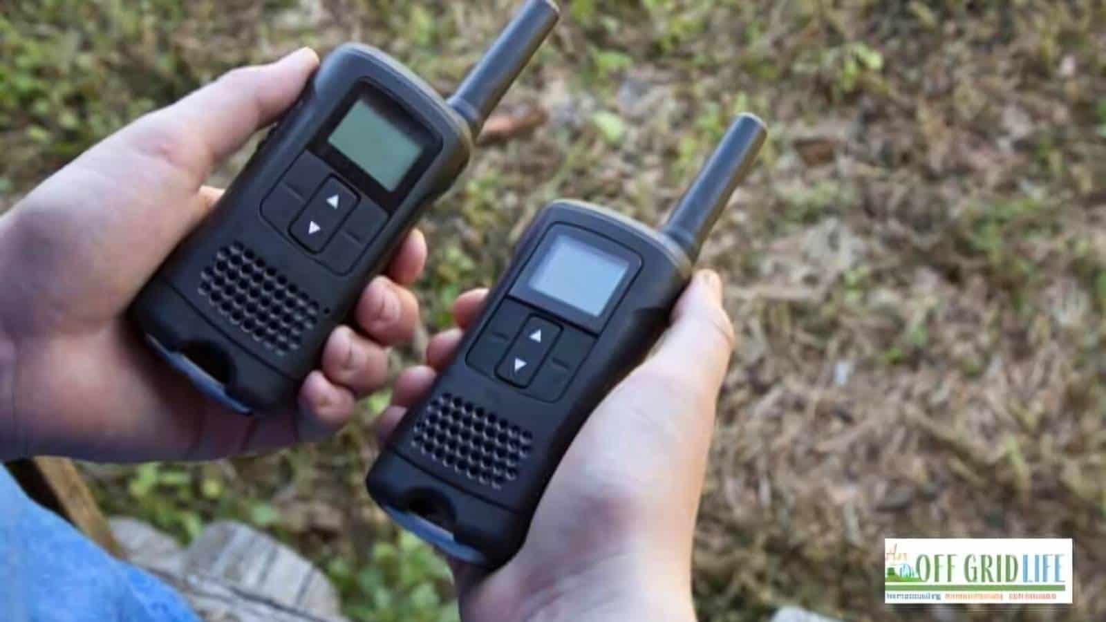 Two hands holding black walkie-talkies with small digital screens and buttons are positioned over a grassy background, capturing the spirit of off-grid living articles.