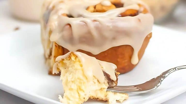 Close-up of a cinnamon roll with icing on a white plate, with a fork holding a bite-sized piece.