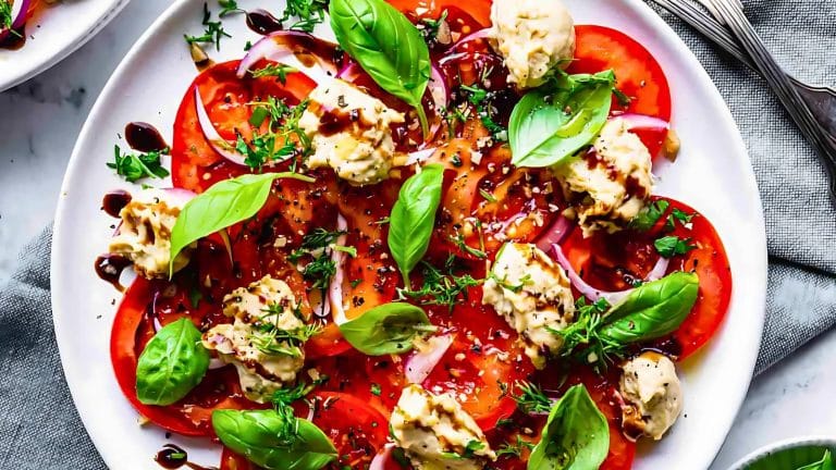 A plate of sliced tomatoes topped with dollops of hummus, fresh basil, red onions, and herbs, drizzled with balsamic glaze.