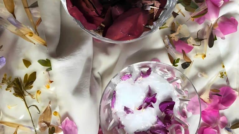 Two glass bowls filled with flower petals, one with ice, rest on a white silky surface surrounded by scattered petals and small flowers.