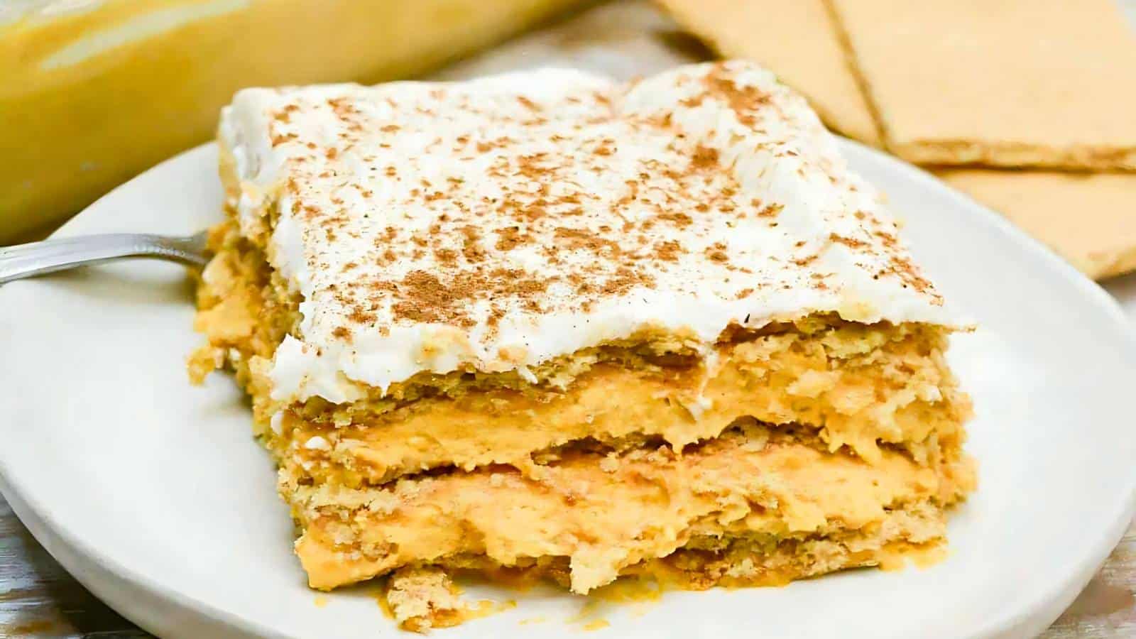 A slice of layered pumpkin dessert with cream topping and a sprinkle of cinnamon, served on a white plate with a fork.