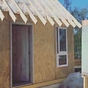 A small, unfinished wooden structure with an open door and window sits under construction outdoors. Surrounding trees and a banner advertising "Off Grid Life" are visible.