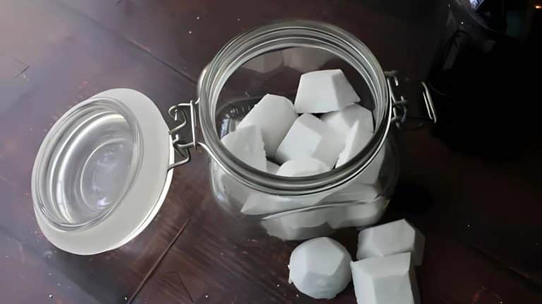 An open glass jar on a wooden table, containing several white, hexagonal sugar cubes with a few scattered outside.