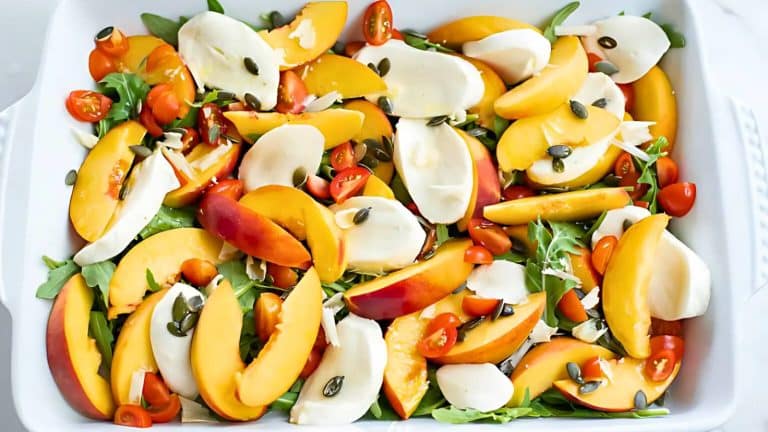 A colorful salad with sliced peaches, mozzarella, cherry tomatoes, arugula, and pumpkin seeds in a white dish.