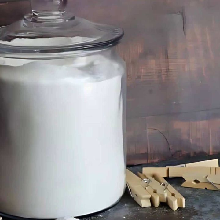 A large glass jar filled with white powder is placed on a dark surface. Several beige wooden clothespins are scattered nearby, against a wooden backdrop.