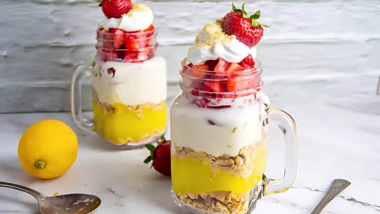 Two mason jar desserts with layers of lemon pudding, crushed cookies, strawberries, and whipped cream, topped with strawberries. A lemon and spoon are nearby on a marble surface.